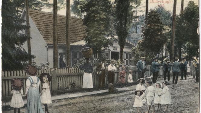Uz ielas redzami cilvēki, fonā aug koki un atrodas balta māja