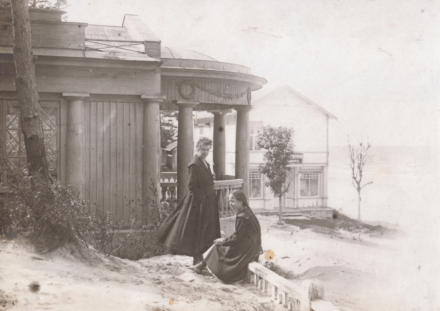 Melnbalta fotogrāfija. Jauna meitene sēž uz zemas koka sētas, blakus stāv sieviete, aiz viņām paviljons – vienstāva koka konstrukciju celtne neoklasicisma arhitektūras stilistikā. Fonā divstāvu ēka un jūra.