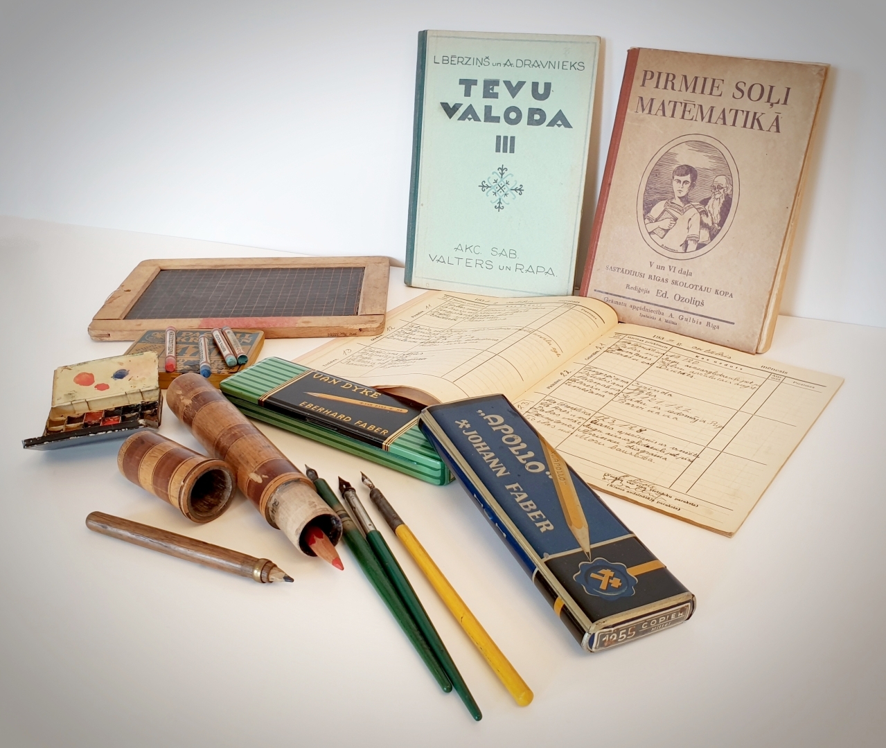 Divas mācību grāmatas, dienasgrāmata, skolēna tāfele koka rāmī un rakstāmpiederumi: zīmuļi, spalvaskāti apaļā koka penālī, divi metāla penāļi, akvareļu krāsas metāla kastītē un pasteļkrītiņi.