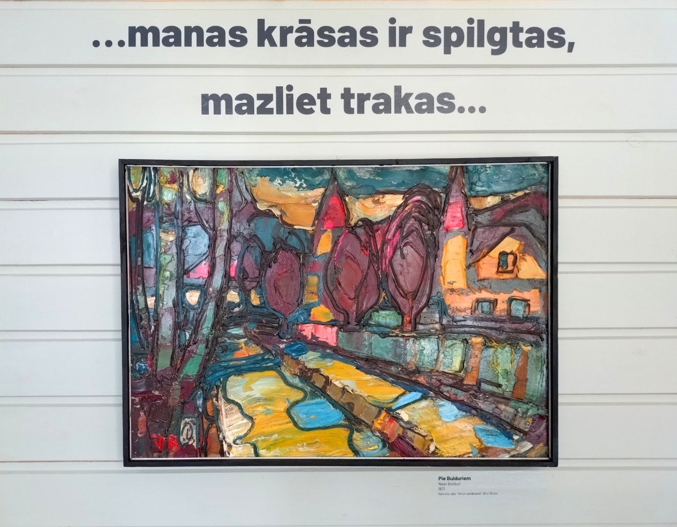 Uz sienas izstādes nosaukums, zem tā glezna spilgtās krāsās