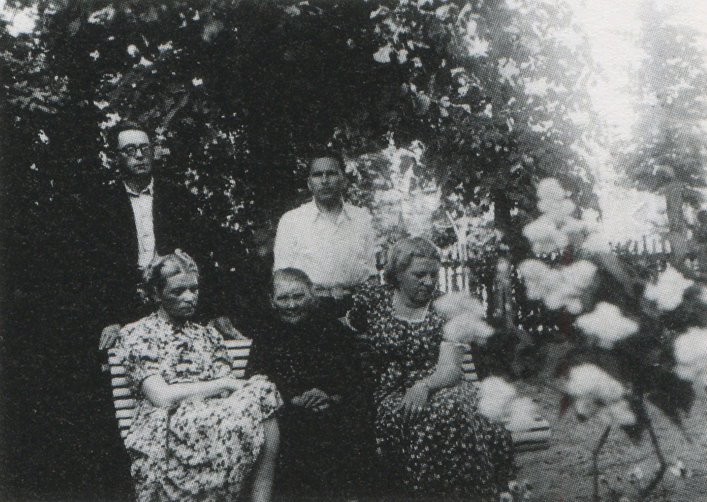 Melnbalta fotogrāfija. Vasaras skats. Trīs sievietes sēž dārzā uz sola, aiz viņām stāv divi vīrieši. 