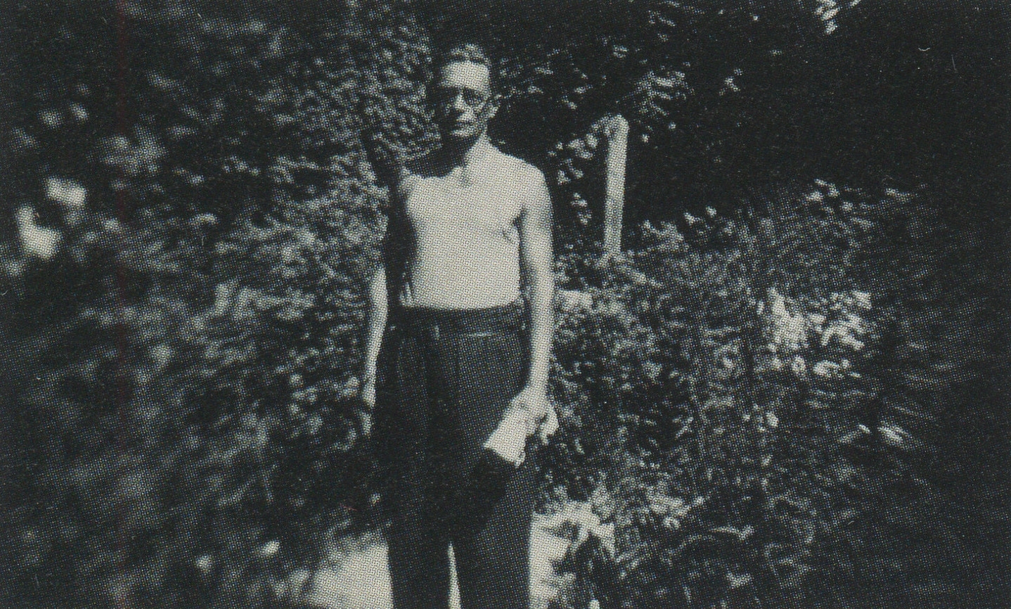 Melnbalta fotogrāfija. Vīrietis vidējos gados ar brillēm, ģērbies baltā bezpiedurkņu kreklā un tumšās biksēs, stāv dārzā.  