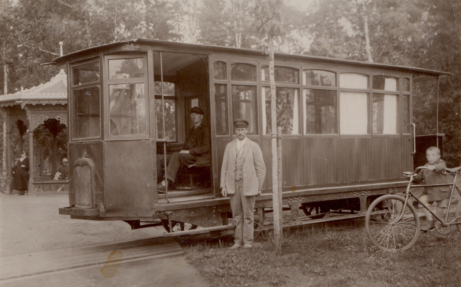 Melnbalta fotogrāfija. Viens tramvaja vagons, kurā sēž vīrietis, pie durvīm stāv vīrietis gaišās drēbēs, labajā malā zēns ar velosipēdu. Fonā cilvēki pie mežģīņrakstu lapenes.