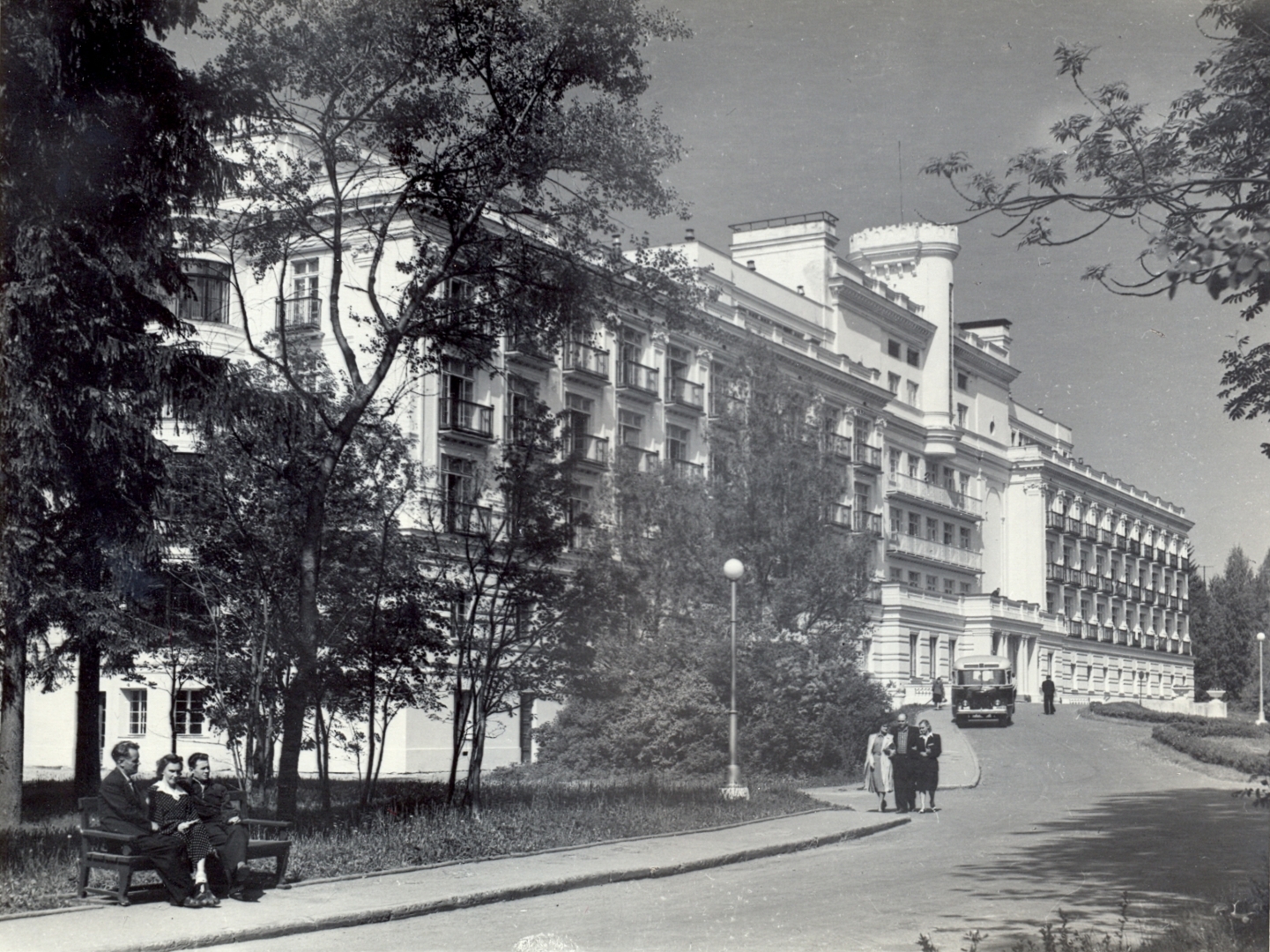 Melnbalta fotogrāfija. Piecstāvu celtne ar jumta terasēm un torni centrā. Celtnes noformējumā bagātīgi izmantoti klasicisma arhitektūras elementi - kolonnas, balustrādes, pilastri, dzegas. Gar ēkas fasādi plats piebraucamais ceļš, pa kuru brauc autobuss, pa ietvi iet cilvēki.