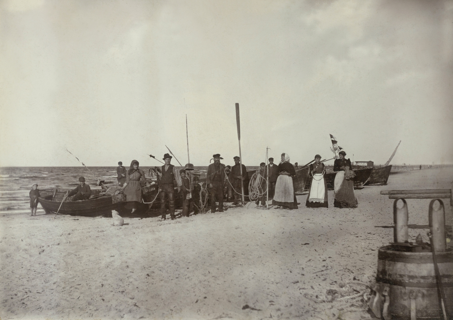 Melnbaltas fotogrāfijas kopija. Pludmales skats ar zvejas laivām, priekšplānā stāv zvejnieki ar airiem, enkuru, virvēm rokās, turpat četras sievietes priekšautos.