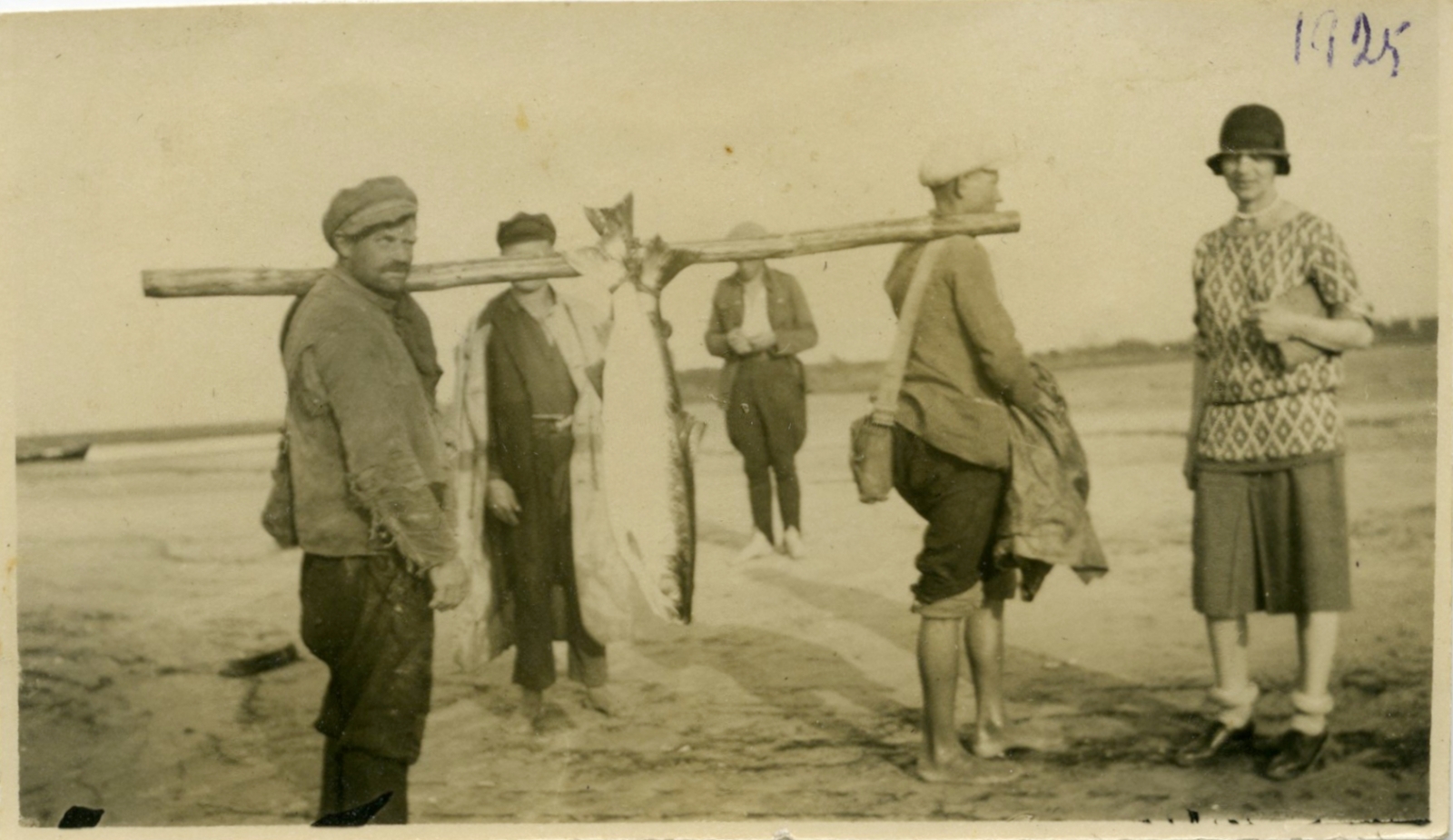 Melnbalta fotogrāfija (sēpija). Divi vīrieši, kas katrs uz viena pleca atbalstījuši apaļu koku, uz kura uzkarinātas divas lielas zivis. Blakus stāv pilsētnieciski ģērbta sieviete, fonā redzami vēl divi vīrieši.