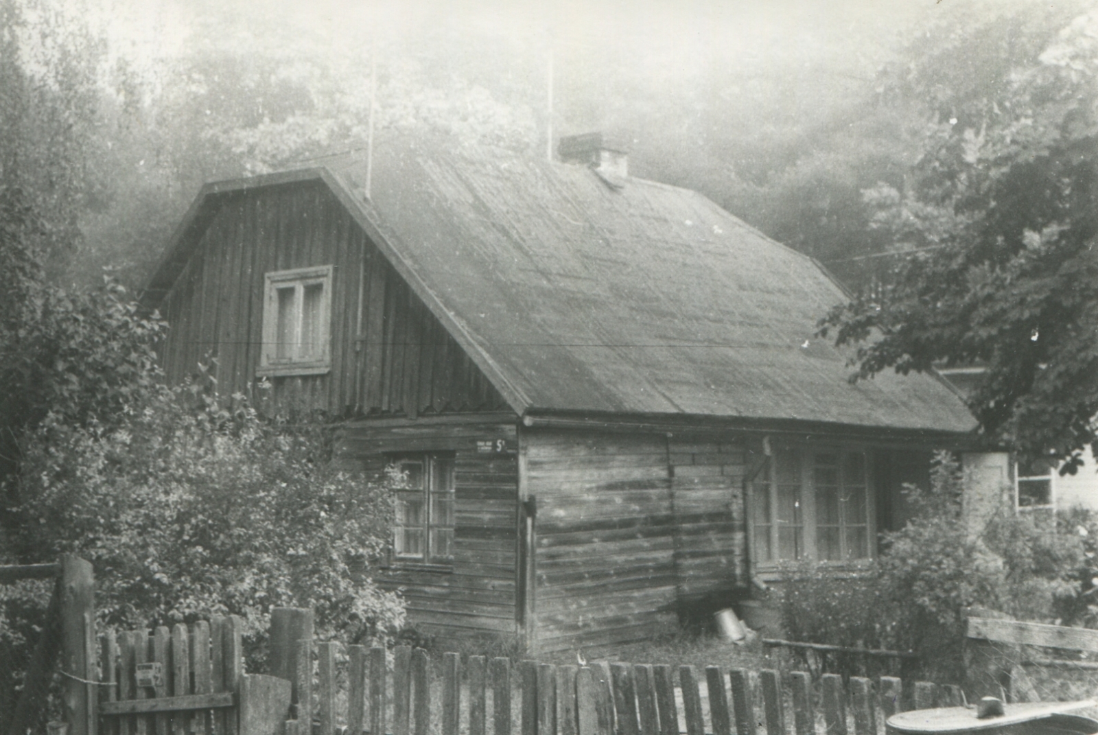 Melnbalta fotogrāfija. Vienstāva koka ēka ar mansarda izbūvi, priekšplānā koka latu žogs.