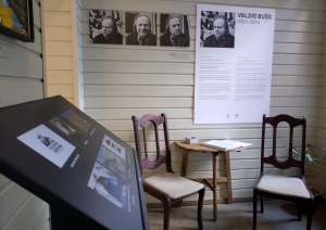 Telpā galds ar diviem krēsliem, pie sienas Valda Buša bildes