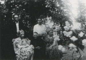Melnbalta fotogrāfija. Vasaras skats. Trīs sievietes sēž dārzā uz sola, aiz viņām stāv divi vīrieši. 