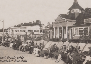 Melnbalta fotoatklātne. Sievietes un vīrieši vasaras drēbēs garā rindā sēž auduma atpūtas krēslos, fonā apbūve – Majoru sporta paviljons ar apaļu torni, tālāk J. Kronberga virpuļgrīdas restorāns – divstāva ēka ar ļoti daudz logiem un kupolveida ieeju.