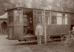 Melnbalta fotogrāfija. Viens tramvaja vagons, kurā sēž vīrietis, pie durvīm stāv vīrietis gaišās drēbēs, labajā malā zēns ar velosipēdu. Fonā cilvēki pie mežģīņrakstu lapenes.