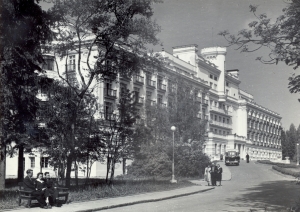Melnbalta fotogrāfija. Piecstāvu celtne ar jumta terasēm un torni centrā. Celtnes noformējumā bagātīgi izmantoti klasicisma arhitektūras elementi - kolonnas, balustrādes, pilastri, dzegas. Gar ēkas fasādi plats piebraucamais ceļš, pa kuru brauc autobuss, pa ietvi iet cilvēki.