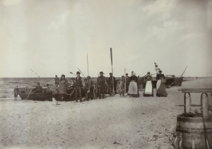 Melnbaltas fotogrāfijas kopija. Pludmales skats ar zvejas laivām, priekšplānā stāv zvejnieki ar airiem, enkuru, virvēm rokās, turpat četras sievietes priekšautos.