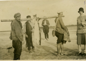 Melnbalta fotogrāfija (sēpija). Divi vīrieši, kas katrs uz viena pleca atbalstījuši apaļu koku, uz kura uzkarinātas divas lielas zivis. Blakus stāv pilsētnieciski ģērbta sieviete, fonā redzami vēl divi vīrieši.