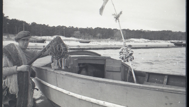 Melnbalts fotonegatīvs. Vīrietis ar tīklu stāv ūdenī pie zvejas laivas.